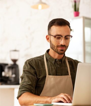 Man in apron working on laptop in coffee shop, developing business strategy for commercial finance solutions.
